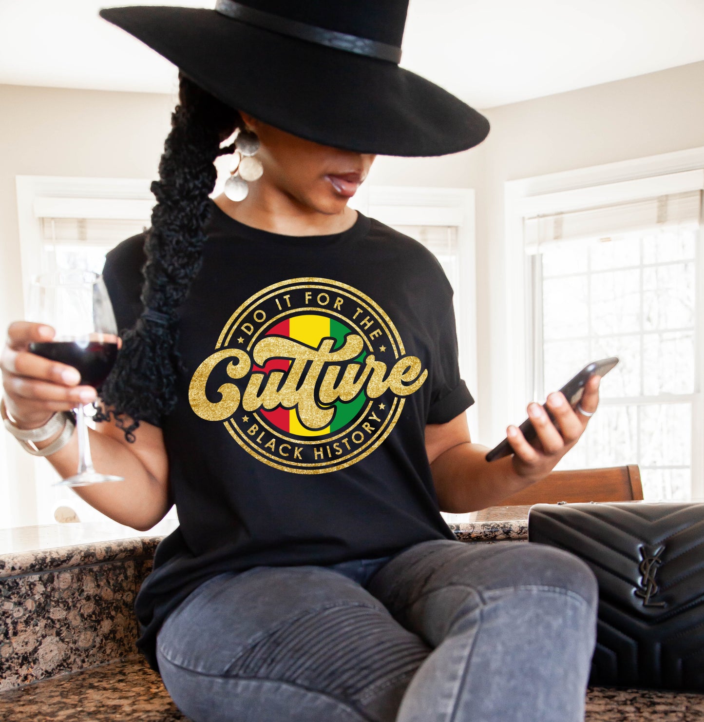 Woman wearing a black t-shirt with a colorful graphic design that says Do it for the culture black history, sitting indoors holding a glass and phone.