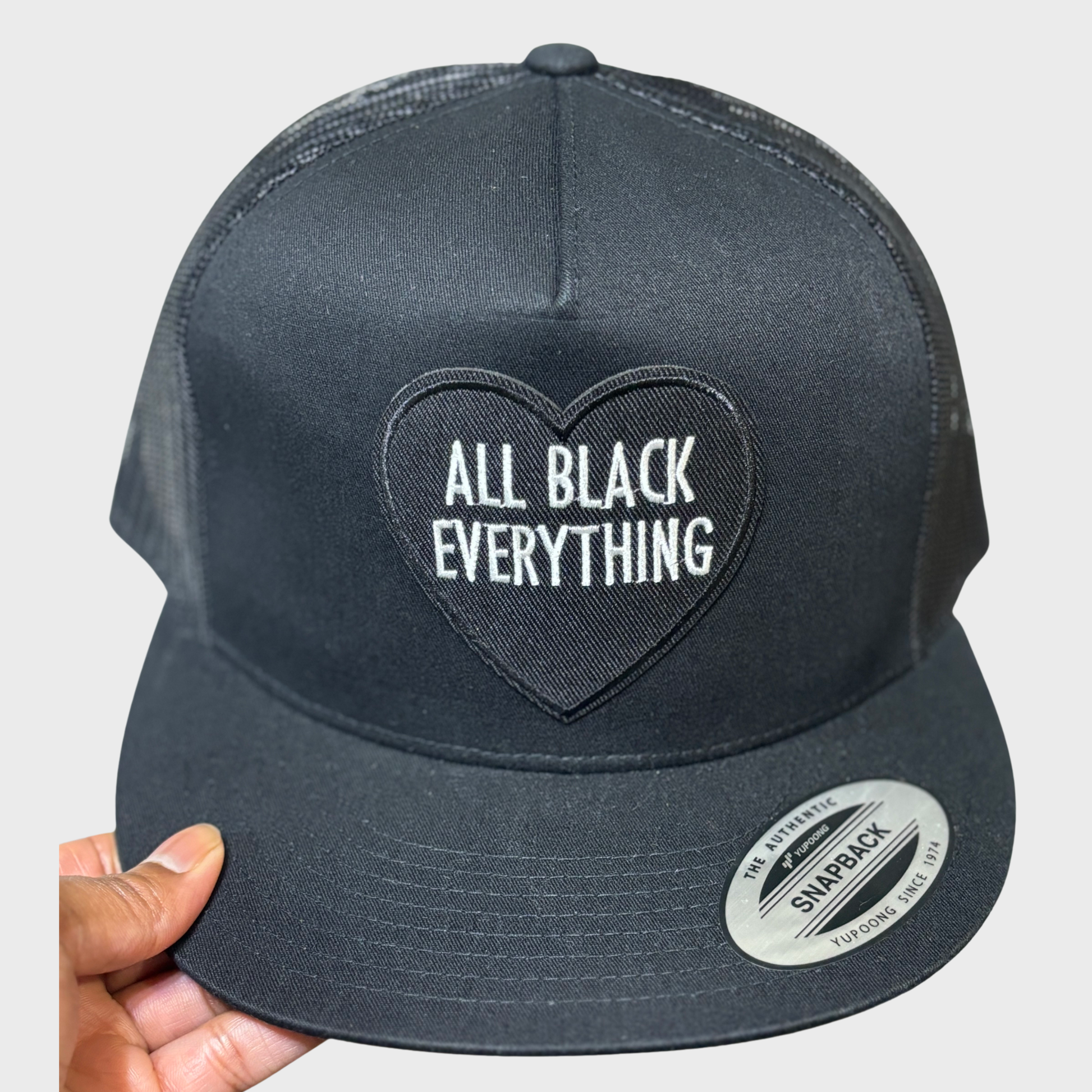 Black snapback hat with a black heart and white text inside the heart that reads "All Black Everything"