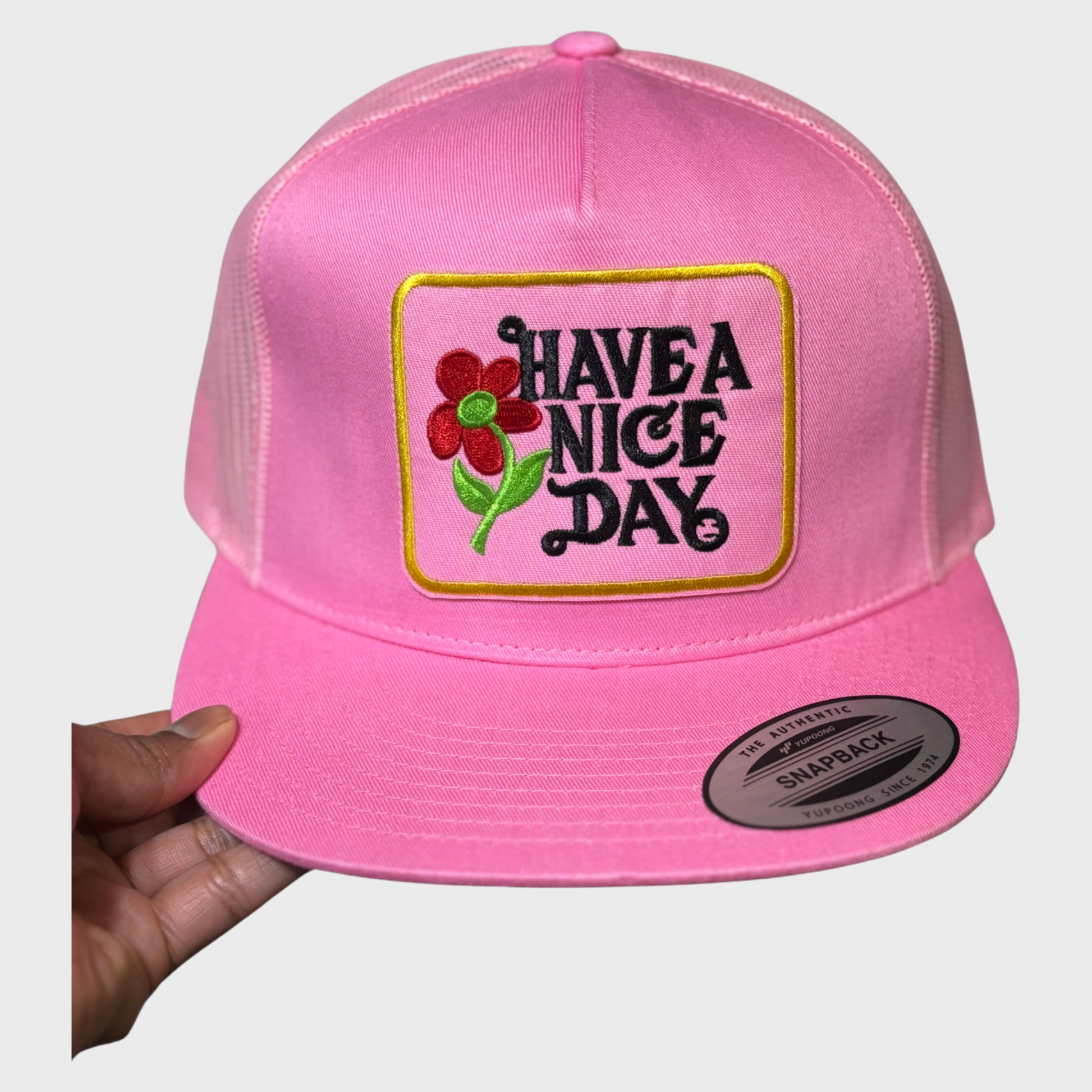 Pink snapback hat with a pink and gold embroidered patch with the text "Have A Nice Day" in black and a red and green flower next to the words